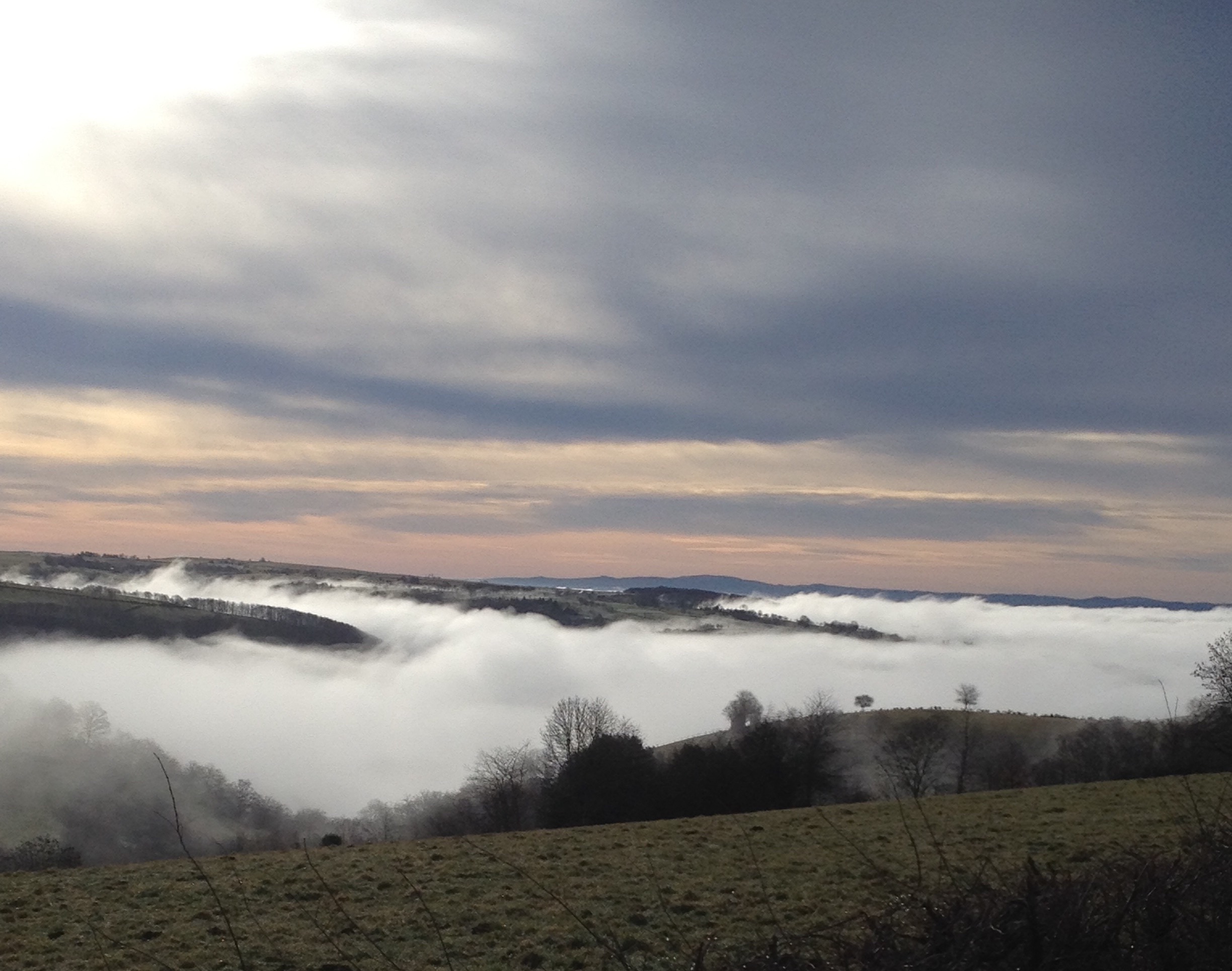 Vallée brumeuse avec de bas nuages entre des collines, ciel doux rose et gris au coucher du soleil